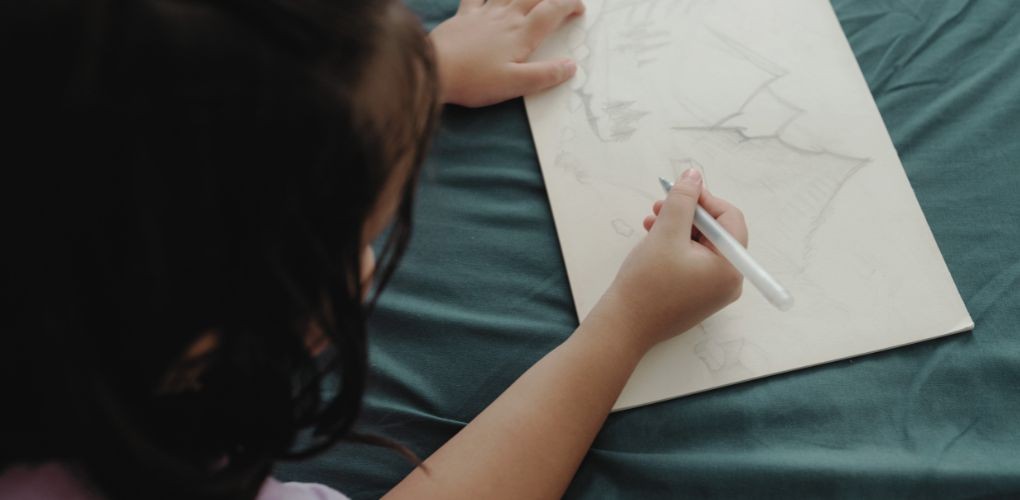 Photo d'une petite fille vue en plongée qui dessine des montagnes sur une feuille blanche.