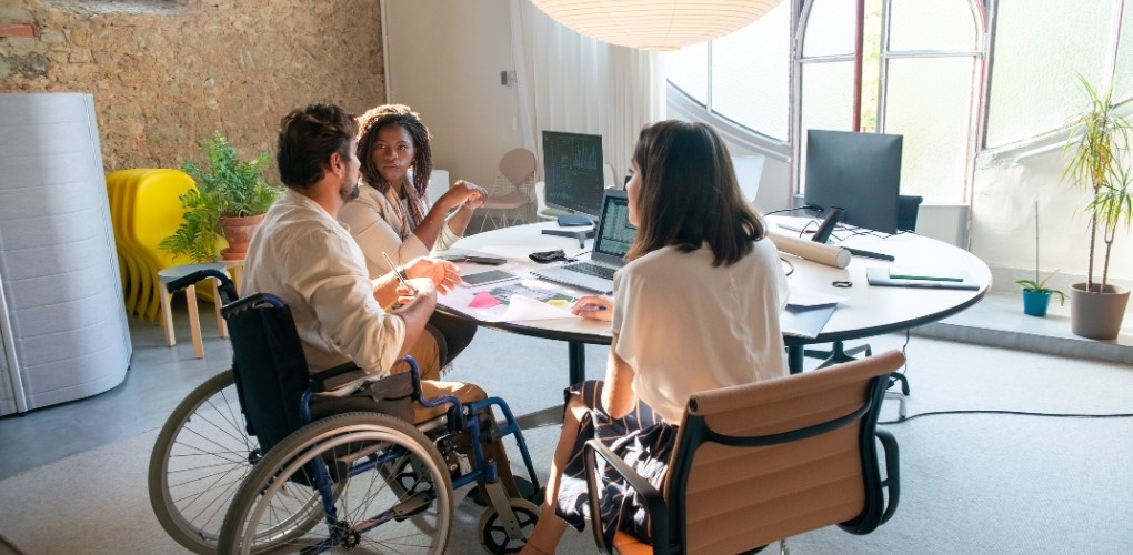 Trois personnes autour d'une table de travail, en discussion devant des ordinateurs, l'homme est en fauteuil roulant.