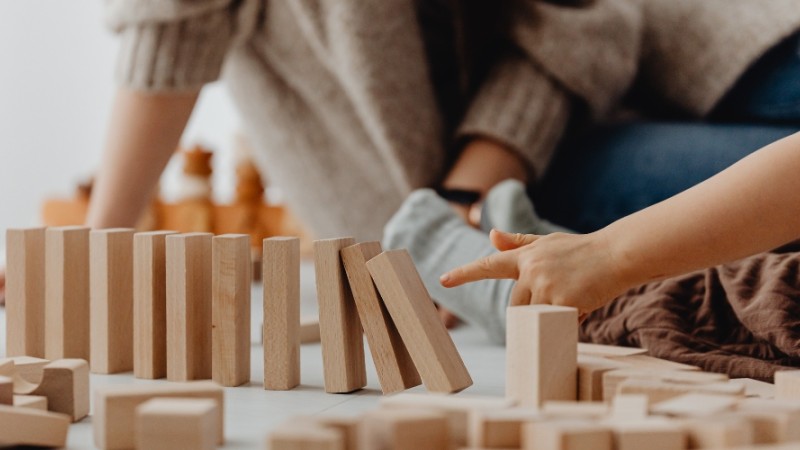 La main d'un enfant qui s'apprête à faire s'écrouler une enfilade de blocs en bois, avec sa mère en arrière-plan.