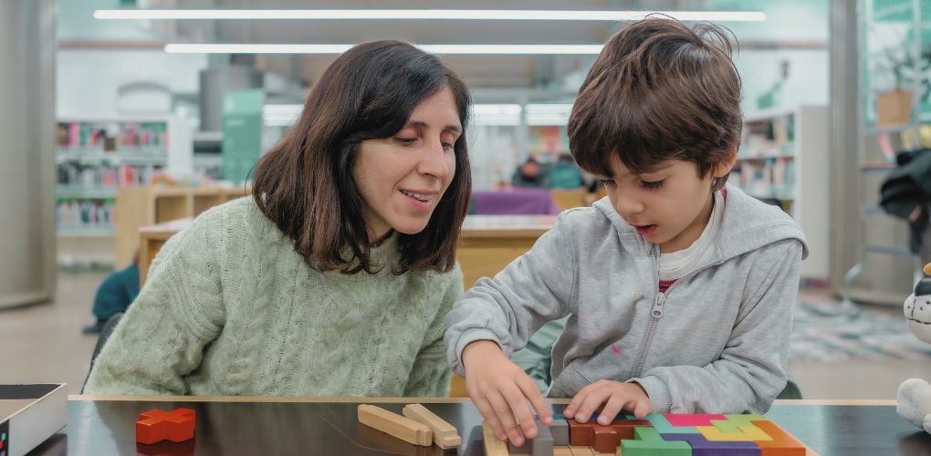 Une mère regarde son enfant assise à côté d'elle qui joue avec des blocs dans une bibliothèque.