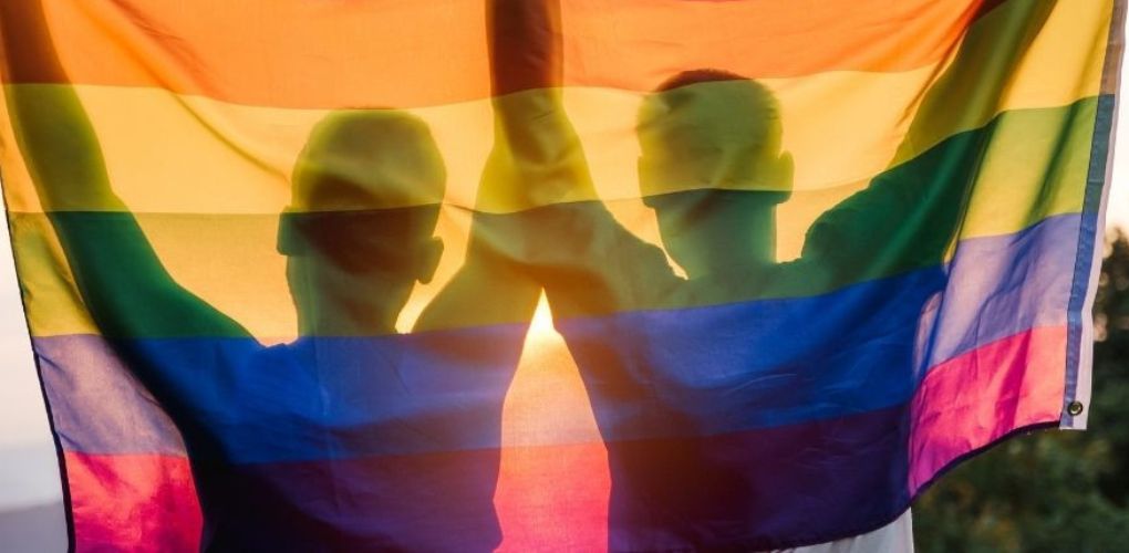 Le drapeau arc-en-ciel tenu par deux personnes dont on voit les silhouettes à travers le drapeau.