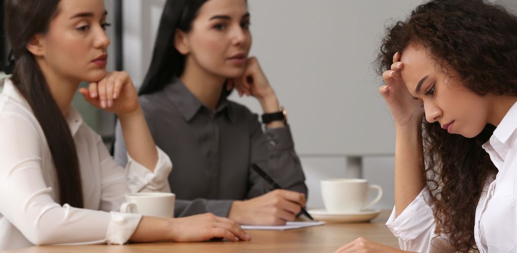 Trois femmes attablées dans un environnement professionnel,. Deux d'entre elles regarde la troisième qui a l'air préoccupé.